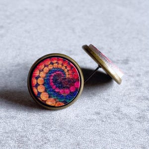 May include: A pair of round stud earrings with a colorful tie-dye design. The earrings feature a bronze-colored metal frame and post. The tie-dye pattern includes shades of orange, pink, blue, and purple, creating a vibrant and eye-catching accessory.