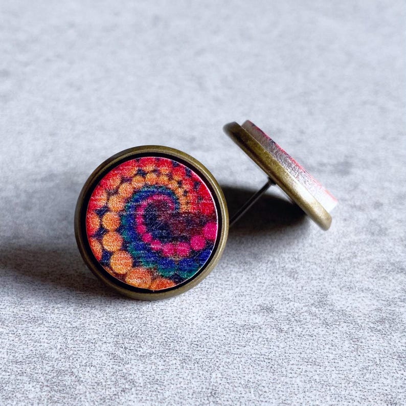 May include: A pair of round stud earrings with a colorful tie-dye design. The earrings feature a bronze-colored metal frame and post. The tie-dye pattern includes shades of orange, pink, blue, and purple, creating a vibrant and eye-catching accessory.