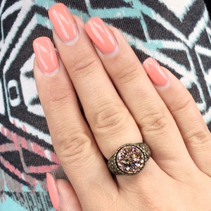 Peut inclure: Une main avec un vernis &agrave; ongles de couleur p&ecirc;che porte une bague avec une grosse pierre ronde rose iris&eacute;e sertie dans une bande de m&eacute;tal bronze.