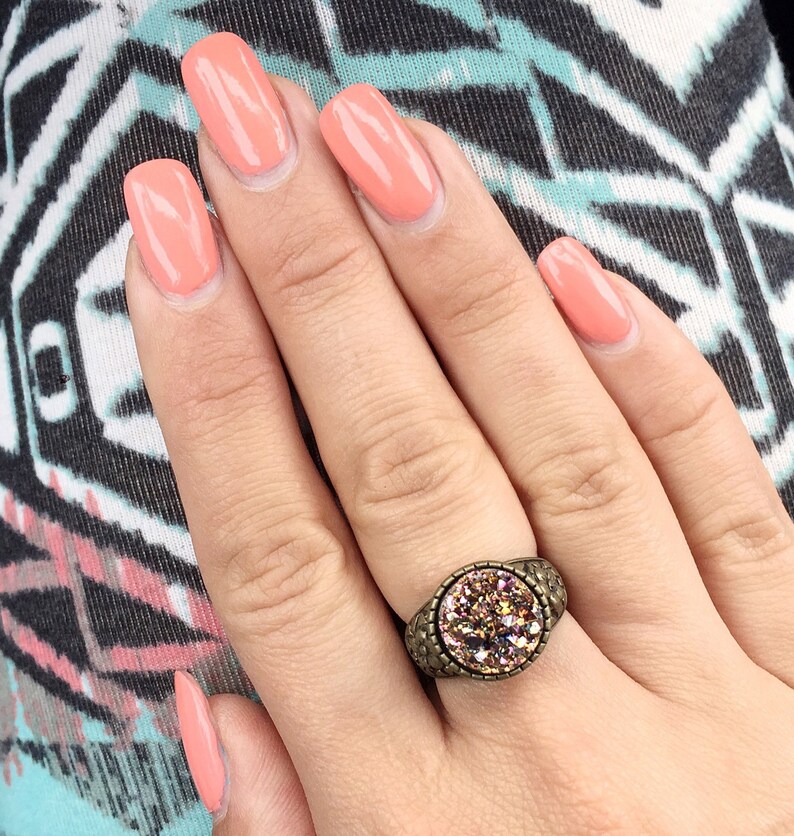 Peut inclure: Une main avec un vernis &agrave; ongles de couleur p&ecirc;che porte une bague avec une grosse pierre ronde rose iris&eacute;e sertie dans une bande de m&eacute;tal bronze.