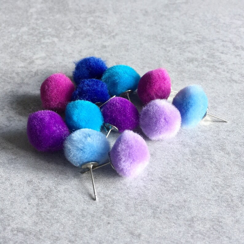 Puede incluir: Una colecci&oacute;n de pompones coloridos en alfileres, incluyendo morado, azul, rosa y lavanda.