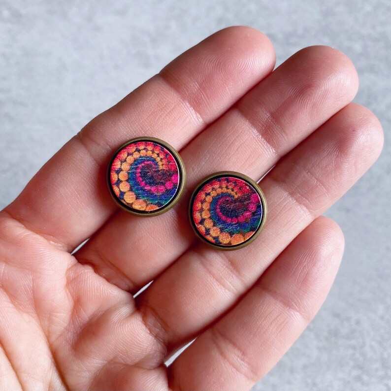 May include: A pair of round stud earrings with a colorful spiral design. The earrings feature a bronze-colored metal frame and a vibrant pattern of orange, yellow, blue, and pink. The earrings are small and suitable for everyday wear.