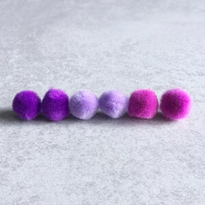Puede incluir: Seis pompones en un degradado de colores morado, lavanda y rosa. Los pompones est&aacute;n dispuestos en una fila sobre una superficie gris claro.