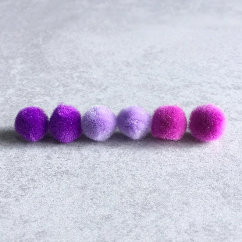 Puede incluir: Seis pompones en un degradado de colores morado, lavanda y rosa. Los pompones est&aacute;n dispuestos en una fila sobre una superficie gris claro.