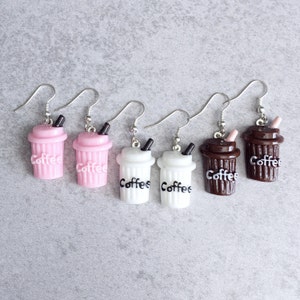 May include: Five coffee cup-shaped dangle earrings in pink, white, and brown. Each earring has a black straw and the word "Coffee" in white. The earrings are suspended from silver-coloured hooks.