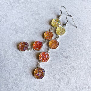 May include: A pair of dangle earrings with seven round, iridescent, glitter-filled charms. The charms are graduated in size, with the largest charm at the bottom and the smallest charm at the top. The charms are connected by silver chains.