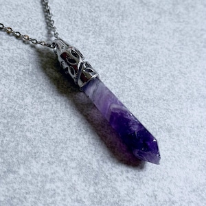 Pode incluir: Um colar prateado com um pingente de cristal de ametista polida. O cristal &eacute; de cor roxa escura com listras mais claras, e o pingente tem uma tampa prateada decorativa. A corrente &eacute; de estilo delicado.