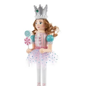 Könnte beinhalten: Dekorativer Nussknacker in Form einer Ballerina. Sie trägt eine silberne Krone, ein rosa-blaues Kleid und Ballettschuhe. Sie hält zwei Lollis. Der Sockel ist weiß. Die Höhe beträgt ca. 25 cm.