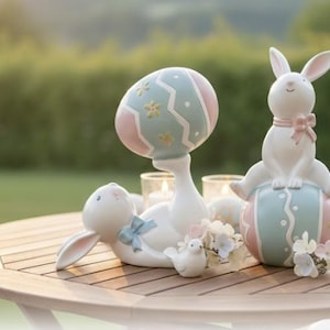 May include: Easter-themed ceramic figurines. A white bunny sits atop a pastel-colored egg, while another bunny reclines. A third figurine is a small white bird. The egg and bunny figures have pink and blue accents. The figurines are on a wooden table.