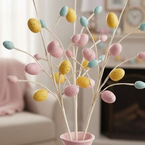 May include: A pink vase holds decorative branches adorned with speckled pastel-colored Easter eggs. The eggs are in shades of yellow, pink, and blue. The branches are white and the vase is a soft pink.