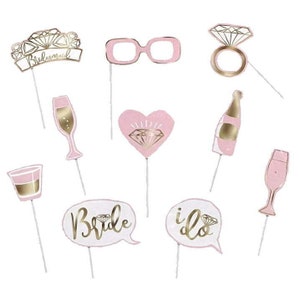 May include: Pink and gold bridal shower photo booth props. The props include a bride and groom speech bubble, a champagne glass, a heart with a diamond, a ring, a bottle of champagne, a shot glass, and a pair of glasses.