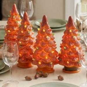 May include: Four conical ceramic trees with a glossy orange finish, each featuring small, multi-colored illuminated bulbs embedded throughout the textured branches. The trees stand on circular bases and are arranged in a group on a table.