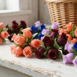 May include: Artificial rose bouquets in shades of peach, burgundy, blue, and purple. The roses have green leaves and are arranged on a distressed white surface. A wicker basket is visible in the background.