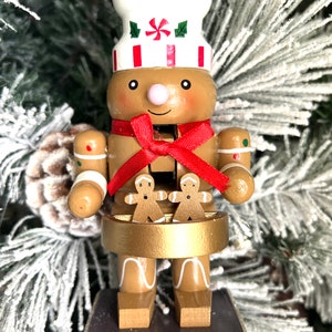 May include: A gingerbread man nutcracker wearing a white chef's hat with red and white stripes, a red bow, and holding two gingerbread cookies.