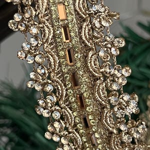 Op de afbeelding: Close-up van een goudkleurige decoratieve rand met ingewikkelde bloemen- en bladerpatronen. De rand is versierd met sprankelende heldere en amberkleurige strass-steentjes. De centrale band heeft rechthoekige amberkleurige stenen.