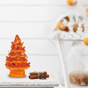 Op de afbeelding: Oranje keramische kerstboom met kleurrijke lichtjes. De boom is versierd met rode, gele en gouden lichtjes. Kaneelstokjes en steranijs zijn met touw samengebonden en naast de boom geplaatst.