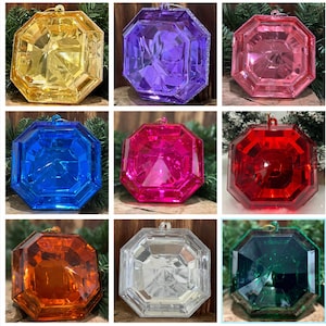 Op de afbeelding: Assortiment van acht achthoekige kerstversieringen. De versieringen zijn gemaakt van helder plastic en hebben een facetgeslepen ontwerp. De versieringen zijn in verschillende kleuren, waaronder rood, blauw, groen, roze, paars, goud, zilver en helder.