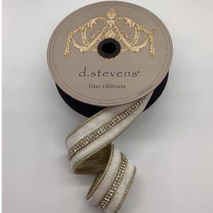 Op de afbeelding: Een spoel met fijne linten van d.stevens. Het lint is wit met een gouden rand en een rij strass-steentjes. De spoel is beige met gouden decoratieve accenten en de tekst "d.stevens fine ribbons".