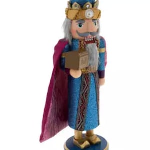 May include: A decorative wooden nutcracker in the form of a king, wearing a blue and gold crown, blue robe, and a purple cape. The nutcracker is holding a small brown box and stands on a blue base.