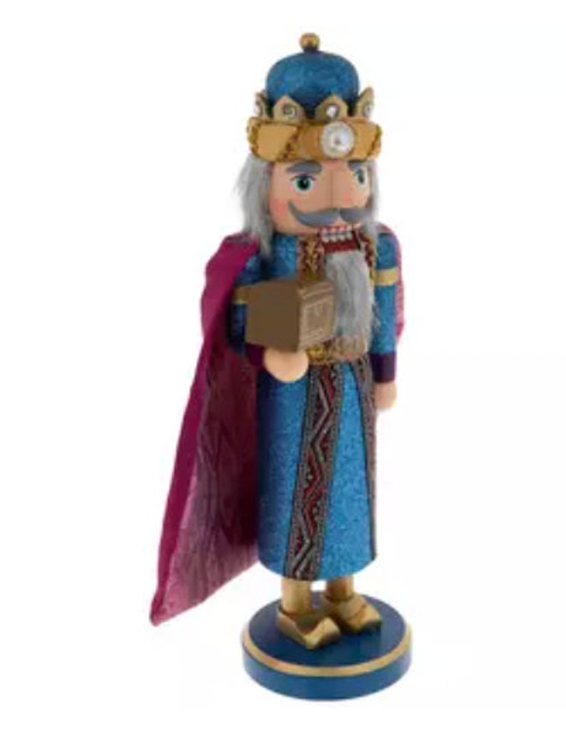 May include: A decorative wooden nutcracker in the form of a king, wearing a blue and gold crown, blue robe, and a purple cape. The nutcracker is holding a small brown box and stands on a blue base.