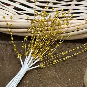 May include: A bundle of decorative floral stems with gold-colored spherical beads. The stems are attached to a white wrapped base. The background includes a woven basket and a brown surface.