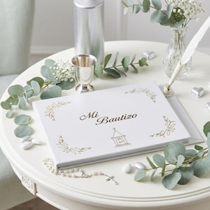 May include: A white book with gold floral accents and the words "Mi Bautizo" is displayed on a white table. A silver chalice, a decorative pen, and a small vase with flowers are also on the table. Eucalyptus and candies surround the book.