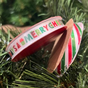 May include: Two spools of Christmas ribbon. One ribbon is white with the words "MERRY CHRISTMAS" in red and green, with gift and tree icons. The other ribbon has red and green stripes. The ribbons are on a green background.