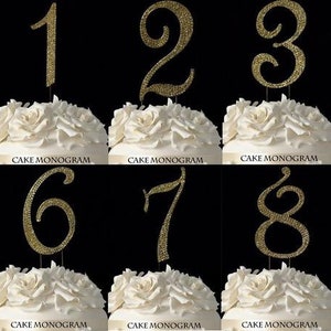 Könnte beinhalten: Zehn goldene Glitzer-Kuchentopper, jeder mit einer anderen Zahl von 0 bis 9. Die Zahlen sind in einer kursivschriftlichen Schriftart und an einem dünnen, klaren Stiel befestigt.