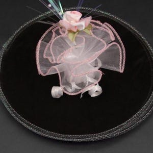 Könnte beinhalten: Ein rundes Tablett aus schwarzem Samt mit einer rosa Blume und einer weißen Tüllschleife darauf.