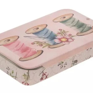 Puede incluir: Una caja de costura rectangular rosa con una tapa que presenta ilustraciones de acuarela de carretes de hilo en rosa, azul y verde. Los lados de la caja tienen un patrón floral, y la tapa tiene un diseño floral y de aguja.