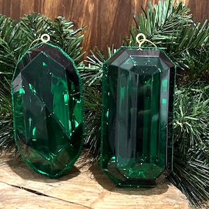 Op de afbeelding: Twee groene glazen kerstversieringen in de vorm van grote, gefacetteerde edelstenen. De versieringen hebben een gouden hanger.