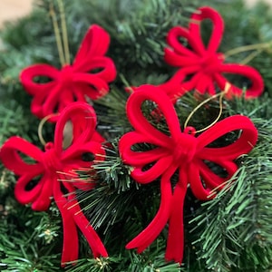 May include: Four bright red velvet bow ornaments with gold hanging strings are displayed on a green Christmas tree. The bows have long, flowing tails and are arranged in a festive display, perfect for holiday decorating.