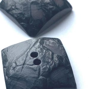 Può includere: Due bottoni quadrati grigio scuro con una superficie strutturata. Il bottone inferiore ha due fori per cucire. I bottoni sono su uno sfondo bianco.