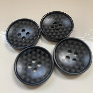 Könnte beinhalten: Vier schwarze Knöpfe mit einem gewebten Karbonfasermuster. Jeder Knopf hat vier Löcher zum Annähen und einen erhöhten Rand. Die Knöpfe sind rund und scheinen aus einem strapazierfähigen Material zu bestehen.