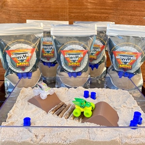 Puede incluir: Un juego de Monster Jam Mini con un recipiente de pl&aacute;stico transparente lleno de arena, una rampa y camiones de juguete. Varias bolsas selladas con el logotipo de Monster Jam est&aacute;n en el fondo. El juego incluye barriles azules y troncos falsos.