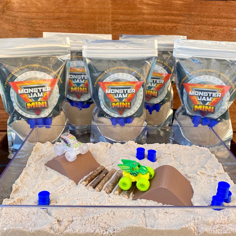 Puede incluir: Un juego de Monster Jam Mini con un recipiente de pl&aacute;stico transparente lleno de arena, una rampa y camiones de juguete. Varias bolsas selladas con el logotipo de Monster Jam est&aacute;n en el fondo. El juego incluye barriles azules y troncos falsos.