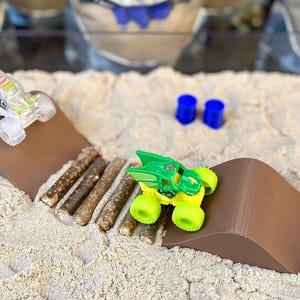 Puede incluir: Una escena de camiones monstruo en miniatura con dos camiones de juguete sobre rampas marrones. Los camiones son verdes y amarillos, y transparentes con acentos coloridos. La escena incluye cilindros azules y una textura arenosa.