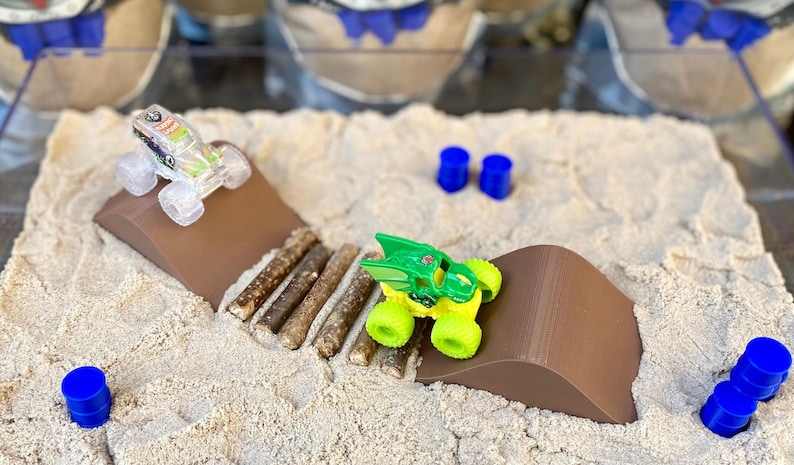 Puede incluir: Una escena de camiones monstruo en miniatura con dos camiones de juguete sobre rampas marrones. Los camiones son verdes y amarillos, y transparentes con acentos coloridos. La escena incluye cilindros azules y una textura arenosa.