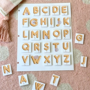 Puede incluir: Un conjunto de fichas de letras del alfabeto con un dise&ntilde;o de globo. Las fichas son blancas con letras naranjas y una cuerda de globo blanca. Las letras est&aacute;n dispuestas en orden alfab&eacute;tico sobre un fondo blanco. Algunas de las fichas est&aacute;n esparcidas sobre una superficie de lunares rosa y blancos.