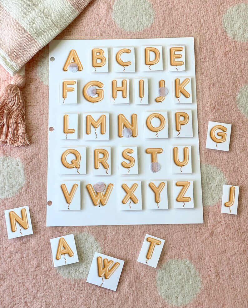 Puede incluir: Un conjunto de fichas de letras del alfabeto con un dise&ntilde;o de globo. Las fichas son blancas con letras naranjas y una cuerda de globo blanca. Las letras est&aacute;n dispuestas en orden alfab&eacute;tico sobre un fondo blanco. Algunas de las fichas est&aacute;n esparcidas sobre una superficie de lunares rosa y blancos.