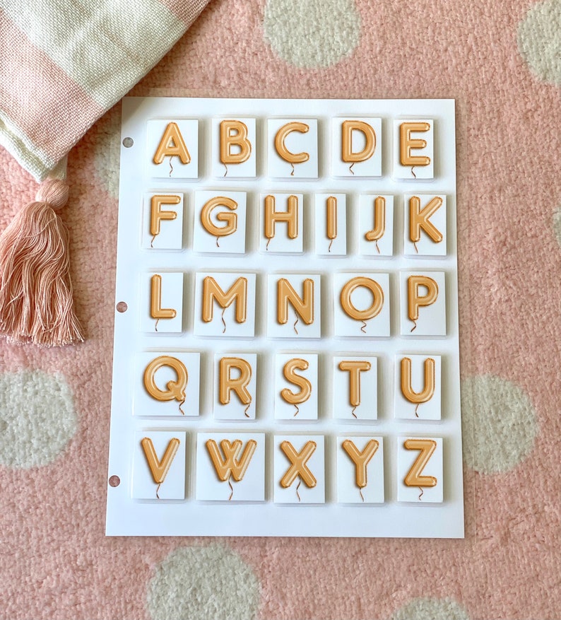 Puede incluir: Pizarra blanca con el alfabeto A-Z en letras de globo doradas. Cada letra est&aacute; en su propio cuadrado.