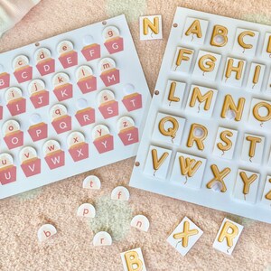 Puede incluir: Dos juegos de fichas de letras del alfabeto. Un juego presenta fichas en forma de cupcake rosa y blanco con las letras A-Z. El otro juego presenta fichas blancas con letras doradas A-Z en forma de globo.