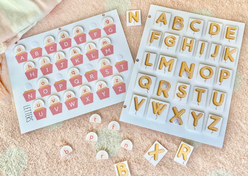 Puede incluir: Dos juegos de fichas de letras del alfabeto. Un juego presenta fichas en forma de cupcake rosa y blanco con las letras A-Z. El otro juego presenta fichas blancas con letras doradas A-Z en forma de globo.