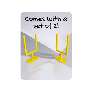 May include: Two yellow miniature football goal posts. The goal posts are on a white and grey background. The text "Comes with a set of 2!" is written in black above the goal posts.