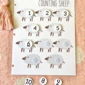 Pu&ograve; includere: Gioco educativo per contare le pecore con illustrazioni di pecore bianche e cerchi bianchi con i numeri da 1 a 10. I numeri sono su uno sfondo bianco con le parole "Counting Sheep" in alto.