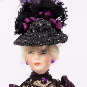 May include: A miniature doll in a purple dress with black lace sleeves and trim. The doll wears a black hat adorned with purple feathers and a matching bow. The doll has blonde hair and purple earrings.