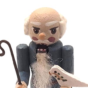 Op de afbeelding: Houten beeldje van een oude man met een lange witte baard, die een staf en een kleine vogel vasthoudt. De man draagt een blauwe jas en heeft een strenge uitdrukking. Het beeldje staat op een kleine houten basis.