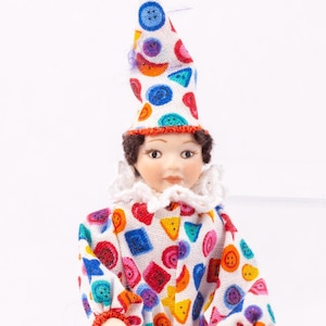 May include: A porcelain doll dressed as a clown. The doll is wearing a white jumpsuit with colorful geometric shapes and buttons. The doll has a red and white hat with a yellow pom-pom on top.
