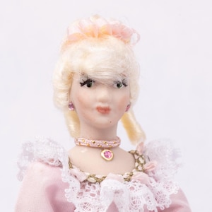 May include: A porcelain doll in a pink dress with white lace trim. The doll has blonde hair styled in an updo with a pink bow. She wears a pink choker necklace and matching earrings. A ring with a pink stone is on her finger.