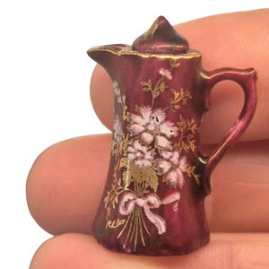 Puede incluir: Jarra de cerámica burdeos en miniatura con un diseño floral en blanco y dorado. La jarra tiene un asa y un pico, y está decorada con un lazo rosa. Un pequeño objeto decorativo.